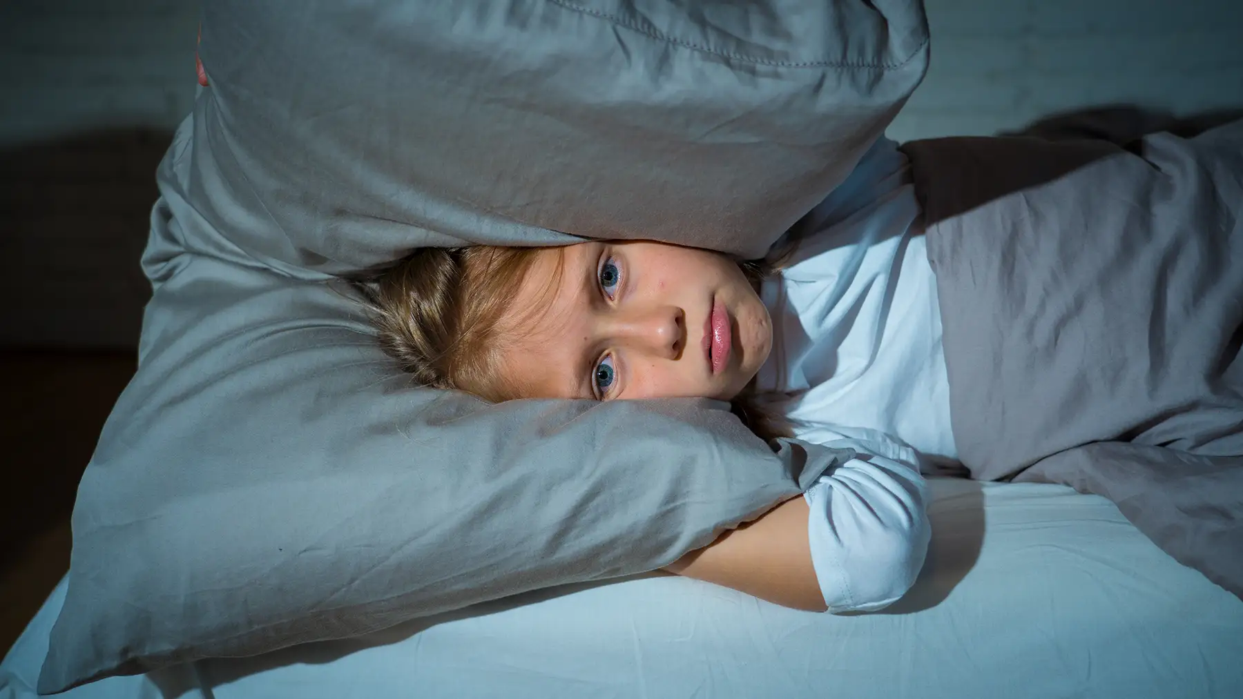 Photo of a girl having trouble sleeping at night holding pillow covering her head and ears, upset