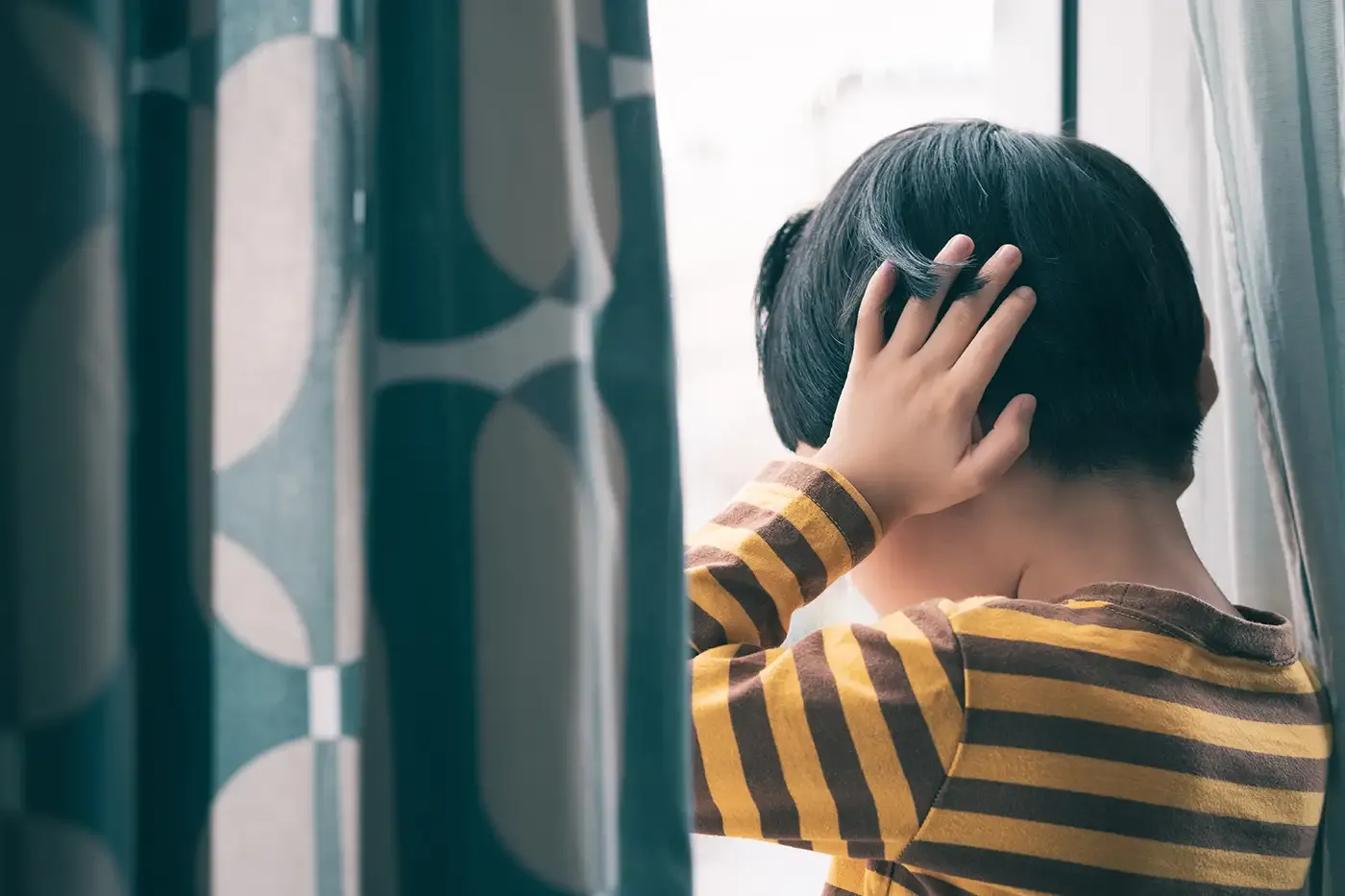 Photo of a boy standing by the window, covering his ears.